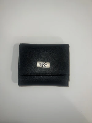 Calvin Klein plånbok - Stilren svart plånbok från Calvin Klein i skinn med metall logga på framsidan. Plånboken har flera fack för kort och sedlar, samt en praktisk knappstängning. Perfekt storlek för att ha med sig överallt och håller dina kort organiserade.
