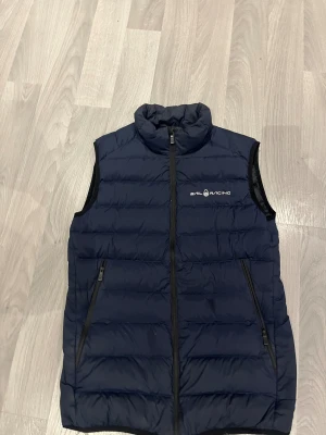 Marinblå dunväst från Sail Racing - Snygg marinblå dunväst från Sail Racing med horisontella quiltade paneler och hög krage. Västen har två dragkedjeförsedda fickor framtill och logotyp på bröstet samt ryggen. Perfekt att slänga över en hoodie för extra värme. Knappt använd, köpte för ca 1,5 år sen, använd max 5 gånger så väldigt fint skick. Nypris 1800kr