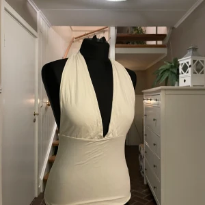 Vit omlott halterneck topp - Säljer en vit halterneck topp med omlott-design och djup v-ringning framtill. Toppen är ärmlös och knyts i nacken för en snygg passform. Materialet känns mjukt och stretchigt, perfekt för att skapa en clean och fräsch look.