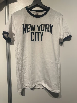 Vit t-shirt New York City Fruit of the Loom - Vit t-shirt från Fruit of the Loom med svart text 'New York City' på bröstet. Svarta kontrastdetaljer vid krage och ärmslut. Liten i storlek. Obs: Alla plagg säljs endast fram till 28/12 
