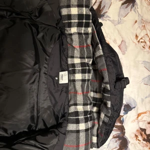 Svart Burberry pufferjacka med huva - Säljer en svart pufferjacka från Burberry med ikonisk rutig insida i svart, vit och röd. Jackan har huva, dragkedja och tryckknappar framtill samt en patch med Burberry London England på ärmen. Perfekt för kalla dagar och har en clean, modern look. Den kan även göras till en väst under varmare dagar. Den var tyvärr för liten vilket betyder att jag behöver sälja den