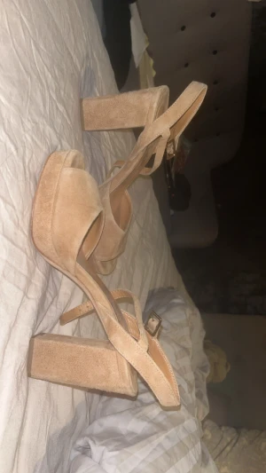 Beige höga sandaler från WERA - Snygga höga sandaler från WERA i beige mocka med bred klack och plattform. Skorna har öppen tå, justerbart spänne och mjuka remmar som korsar foten. Perfekta för dig som gillar stilrena och trendiga skor med extra höjd. Klacken är 9,5 cm hög.