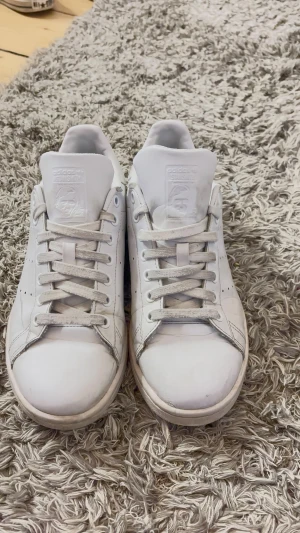 Adidas Stan Smith vita sneakers - Klassiska Adidas Stan Smith sneakers i helvitt skinn med rund tå och platt sula. Skorna har snörning och diskret perforering på sidorna. Stan Smith-logga på plösen och hälen. Perfekt för dig som gillar clean och tidlös stil. Jag kommer rengöra dem så att de nästan ser ut som nya! 