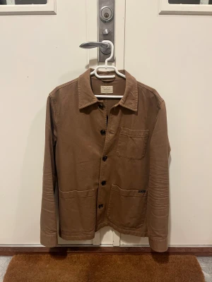 Brun overshirt från Nudie Jeans Co M - Snygg brun overshirt från Nudie Jeans Co i 100% ekologisk bomull. Jackan har klassisk krage, knäppning framtill med svarta knappar och tre praktiska fickor. Perfekt lager-på-lager-plagg med stilren och enkel design.
