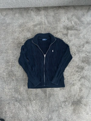 Stickad Ralph lauren full zip - Mörkblå stickad kofta från Polo Ralph Lauren med dragkedja framtill och klassiskt kabelstickat mönster. Tröjan har hög krage och broderad logga på bröstet. Perfekt för lager-på-lager och snygg till jeans eller chinos.