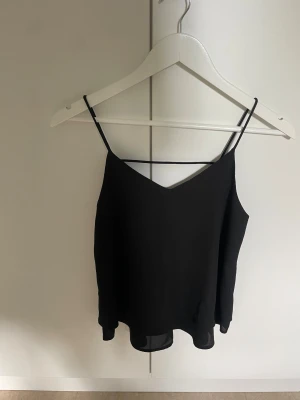 Svart linne med smala axelband - Säljer en svart linnetopp med smala axelband och v-ringad hals. Toppen har en luftig passform och ett extra band över ryggen som ger en snygg detalj. Perfekt för varma dagar och enkel att matcha med shorts eller jeans. Aldrig använd, från veromoda 