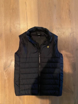 Svart pufferväst från Lyle & Scott - Svart pufferväst från Lyle & Scott Junior med gul logga på bröstet. Västen har ståkrage och dragkedja framtill. Perfekt för lager-på-lager och har en stilren, sportig look som funkar till många outfits. Litet hack i dragkedjan som inte påverkar knäppningen. Därav det otroliga priset 