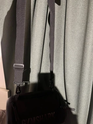 Svart axelväska från Gymshark - Svart axelväska från Gymshark med cylinderformad design och justerbar axelrem. Väskan har dragkedja och ett stilrent Gymshark-tryck på sidan. Materialet är slitstark syntet och botten har Gymshark-logga i relief. Perfekt för gymmet eller när du vill ha med dig det viktigaste.