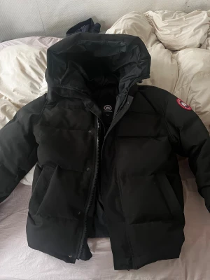 Svart pufferjacka från Canada Goose XS - Svart pufferjacka från Canada Goose i storlek XS. Jackan har en stor huva, dragkedja framtill och klassisk logga på ärmen. Fylld med dun och har rejäla fickor. Perfekt för kalla vinterdagar och har en clean, modern look.