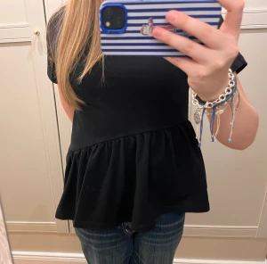 Svart volangblus från COS - Svart tröja från COS i storlek XS. Modellen har korta ärmar, rund hals och en bred volang nedtill som ger en snygg och flowy siluett. Materialet är mjukt och känns som bomull. 