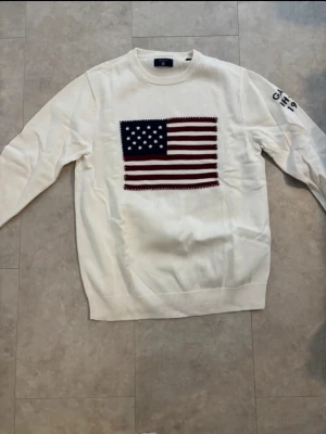 Vit stickad tröja från GANT - Vit stickad tröja från GANT med amerikansk flagga broderad på bröstet och blå text 'GANT NH-CT 1949' på ena ärmen. Tröjan har rund hals och ribbade muddar vid ärmslut och nederkant. Perfekt för dig som gillar klassisk college-stil.