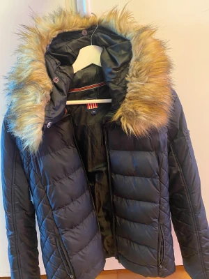 Svart pufferjacka med fuskpäls från Vinson - Svart pufferjacka från Vinson med stor huva och fluffig fuskpäls i beige/bruna toner. Jackan har quiltad design, dragkedja framtill och sidofickor med dragkedja. Perfekt för kalla dagar och har en riktigt snygg streetstil. Jag har två stycken.