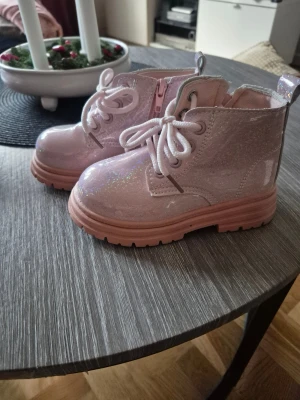 Glittriga rosa boots med snörning - Säljer ett par snygga boots i ljusrosa med glittrig finish och grov platt sula. Skorna har rund tå, snörning framtill och dragkedja på sidan för enkel påtagning. Perfekt för dig som vill sticka ut med lite extra glitter i vardagen.