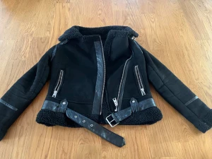 Svart teddyjacka från Zara med bälte - Svart teddyjacka från Zara med mjukt foder, detaljer i skinnimitation och två dragkedjeförsedda fickor. Jackan har bälte i midjan, bred krage och långa ärmar med skinnband. Perfekt för kalla dagar och har en cool bikerstil.