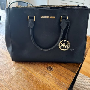 Svart handväska från Michael Kors - Säljer en klassisk svart handväska från Michael Kors med guldfärgade detaljer och logotyp. Väskan har två handtag, dragkedja upptill och flera fack inuti för smart förvaring. En liten vit fläck framtill, se bild. Ca 34 x 24 cm. 