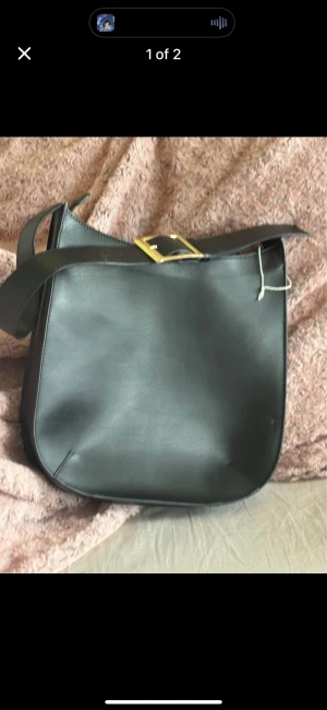 Svart axelväska i skinn med spänne - Stilren svart axelväska i skinn med en bred axelrem och snyggt guldfärgat spänne. Väskan har en enkel, rundad form och är perfekt för dig som gillar minimalistisk design. Passar till många olika outfits och har plats för det viktigaste.