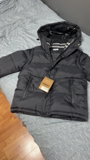 Burberry jacka - Snygg svart pufferjacka från Burberry med huva och dragkedja framtill. Jackan har två framfickor med dragkedja och en stor Burberry London England-patch på ärmen. Insidan har ett randigt foder i grått och vitt. Perfekt för kalla dagar.