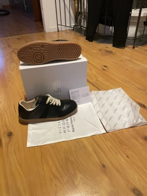 Maison Margiela svarta sneakers - Säljer ett par stilrena gats från Maison margiela! Skorna är i mycket bra skick och knappt använda, 10/10 skick. Enda defekten är på boxen som är lite buktig. Storlek 43. Allting på bilderna tillkommer. Om ni har några frågor så är det bara att höra av er!