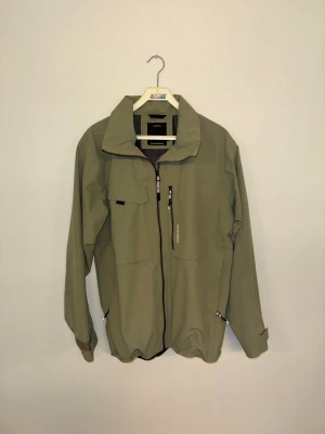 Grön vindjacka från Didriksons - Snygg grön vindjacka från Didriksons med vattenavvisande material och meshfoder. Jackan har flera praktiska fickor med dragkedja, hög krage och justerbara ärmslut. Perfekt för blåsiga dagar och outdoor-aktiviteter. Stilsäker och funktionell design.