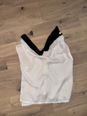 Vit blus med svart krage  - Säljer en vit blus med en snygg svart krage och knappdetalj. Blusen är ärmlös och har dragkedja i ryggen för enkel påklädning. Materialet känns lätt och luftigt, perfekt för dig som gillar stilrena och klassiska plagg.