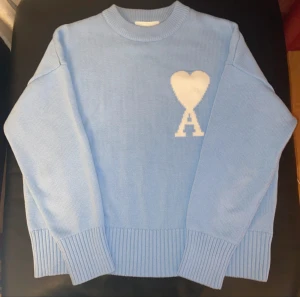 Ljusblå stickad tröja från Ami Paris - Säljer en ljusblå stickad tröja från Ami Paris med ett vitt hjärta och bokstaven A på bröstet. Tröjan har rund hals, långa ärmar och breda ribbade muddar vid ärmslut och nederkant. Perfekt för dig som gillar stilrena detaljer och mjuka material.