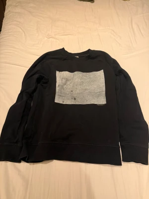 Svart Nudie Jeans Tröja - Svart tröja från Nudie Jeans i 100% ekologisk bomull. Tröjan har ett stort grått tryck på bröstet och klassisk rund hals. Långa ärmar och ribbade muddar vid ärmslut. Perfekt för dig som gillar simpla tröjor och enkel stil.