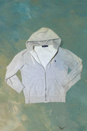 Grå hoodie från Polo Ralph Lauren - Snygg grå hoodie från Polo Ralph Lauren med dragkedja och huva. Tröjan har långa ärmar, snörning vid huvan och klassisk blå Polo-logga broderad på bröstet. Mjuk insida i fleece och praktiska fickor framtill. Perfekt för chill dagar.
