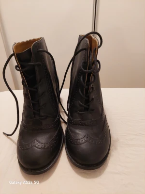 Svarta brogue boots från Ellos, strl 37 - Säljer ett par svarta boots från Ellos i storlek 37. Skorna har klassisk brogue-mönstrad design, snörning framtill och låg klack. Tillverkade i skinnimitation med rund tå och snygga detaljer längs kanterna. Perfekt för dig som gillar stilrena och tidlösa skor.