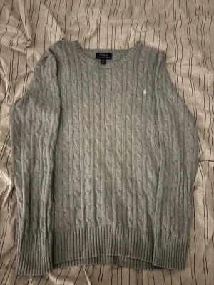 Grå stickad tröja från Polo Ralph Lauren - Snygg grå kabelstickad tröja från Polo Ralph Lauren med rund hals och diskret broderad logga på bröstet. Tröjan har långa ärmar och ribbade muddar vid ärmslut och nederkant. Perfekt för dig som gillar klassisk och stilren look.