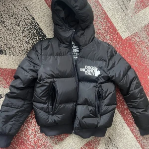 Svart pufferjacka från DSQUARED2 - Säljer en svart pufferjacka från DSQUARED2 med huva och vit logga på bröstet. Jackan har två stora fickor med dragkedja och en rejäl, vadderad look som håller dig varm. Perfekt för dig som gillar streetwear och vill sticka ut med en snygg jacka.