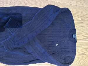 Marinblå stickad tröja Ralph Lauren - Säljer en marinblå stickad tröja från Ralph Lauren med klassiskt kabelstickat mönster och rund hals. Tröjan har långa ärmar och en liten vit broderad logga på bröstet. Är en storlek L (damstorlek)  (passar till en man med S)