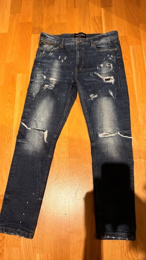 Mörkblå distressed jeans Supply & Demand - Snygga mörkblå jeans från Supply & Demand med slitna detaljer och blekta partier på benen. Modellen har klassisk femficksdesign, raka ben och coola trasiga effekter framtill. Perfekt för dig som gillar en streetstyle-look.