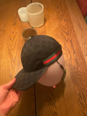 Svart canvas Gucci keps  - Säljer en svart Gucci keps med  GG-mönster över hela kepsen och en snygg grön-röd rand på sidan. Justerbar rem baktill i skinn och böjd skärm. Sömnen är perfekt och taggen är magisk.