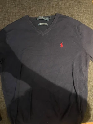 Mörkblå v-ringad tröja Polo Ralph Lauren - Mörkblå v-ringad tröja från Polo Ralph Lauren i 100% pima bomull.  röd  logga på bröstet och ribbade muddar vid hals, ärmslut och nederkant. Säljer då jag växt ur den. Vid eventuella frågor hör av er. MVH Måns 