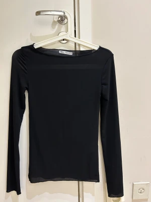  Ny Svart långärmad topp från Zara - Säljer en svart långärmad topp från Zara i storlek S. Toppen har en enkel och stilren design med rund hals och är tillverkad i ett tunt, stretchigt syntetmaterial som sitter snyggt mot kroppen. Perfekt att styla med jeans eller kjol för en clean look.