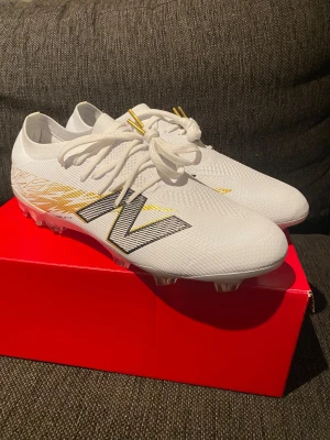 New Balance Furon V8 fotbollsskor 44 - Säljer ett par vita New Balance Furon V8 fotbollsskor med snygga guldfärgade detaljer och mönster på sidan. Skorna har snörning, platt sula med dobbar och är tillverkade i syntetmaterial för lätt känsla på planen. Perfekt för dig som vill sticka ut med stil på matchen.