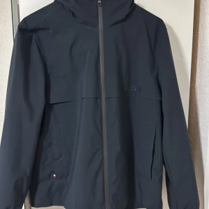 Mörkblå vindjacka Tommy Hilfiger  - Snygg mörkblå vindjacka från Tommy Hilfiger med huva, hel dragkedja och diskret logga på bröstet samt ärmen. Jackan har två fickor framtill och är tillverkad i ett lätt, vattenavvisande material. Storlek M