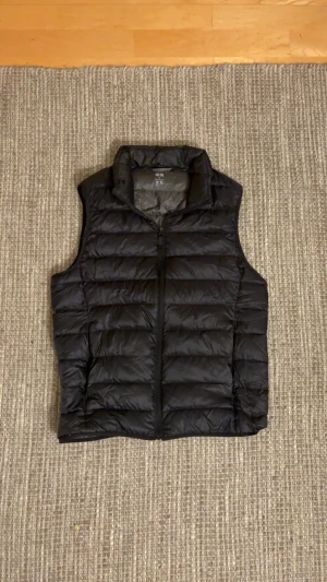 Svart väst från Uniqlo  - Svart, lätt och quiltad dunväst från Uniqlo i storlek XS. Västen har hög krage och dragkedja framtill. Perfekt för lager-på-lager och enkel att bära tack vare sin ultralätta design. det finnd ett jätte litet hål nere pä västen men syns inte