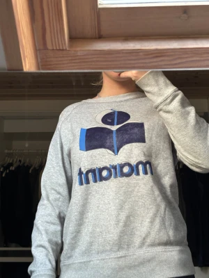 Isabel Marant tröja  - Grå sweatshirt från Isabel Étoile Marant med stort blått och mörkblått logotryck på bröstet. Tröjan har rund hals, ribbade muddar och är tillverkad i mjuk bomull. Perfekt för chill dagar och har en clean, klassisk vibe. Själva märket är lite bortskrapat men annars är tröjan i väldigt bra skick. Självklart äkta🩷🩷