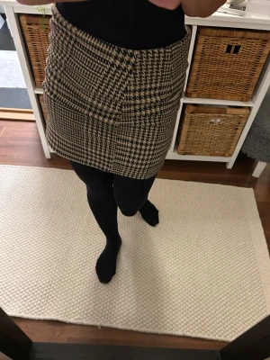 Rutig kortkjol från H&M i beige och svart Stl 32 - Supersnygg kortkjol från H&M med klassiskt rutigt mönster i beige, svart och vitt. Kjolen har rak passform och är tillverkad i ett mjukt material som känns lite som ullmix. Perfekt att styla med strumpbyxor och boots för en trendig look.