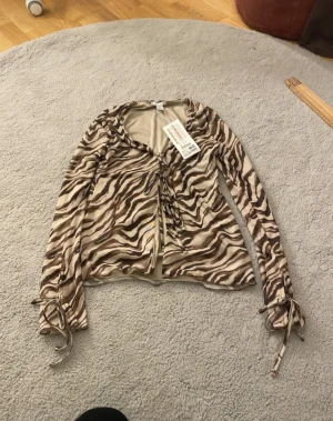 Zebramönstrad långärmad topp från new yorker  - Säljer en snygg långärmad topp från Amisu i storlek XS med zebramönster i beige och bruna toner. Toppen har snörning framtill och vid ärmsluten, samt en v-ringad hals. Materialet är mjuk polyester som känns skönt mot huden.