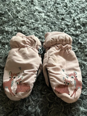 Rosa tumvantar med rådjurstryck från KappAhl - Säljer ett par gulliga rosa tumvantar med sött tryck av rådjur och små hjärtan. Vantarna har ribbade muddar och svart greppvänlig insida i syntetmaterial. Perfekta för kalla dagar och riktigt mysiga att ha på sig.
