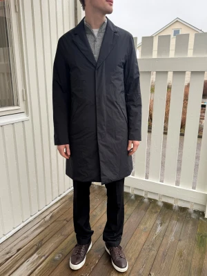 Svart rock från J.Lindeberg - Snygg svart rock från J.Lindeberg med stilren design och klassisk krage. Jackan har en diskret JL-logga på ärmen och är tillverkad i ett slätt syntetmaterial som står emot vind och regn. Passformen är normal och rocken har praktiska sidfickor samt slits bak. Jag Köpte den på J.lindbergs affär i Göteborg och jag har aldrig använt den. 