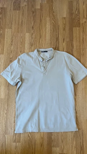 Beige pikétröja från Zara - Snygg beige pikétröja från Zara med korta ärmar och två knappar vid halsen. Tröjan har en klassisk ståkrage och är gjord i mjuk bomull som känns skön mot huden. Perfekt för dig som gillar stilrena och enkla plagg.