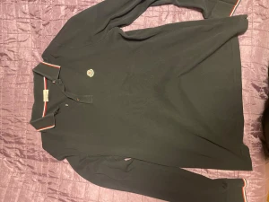 Svart Moncler pikétröja XL - Svart långärmad pikétröja från Moncler i storlek XL. Tröjan har klassisk krage med röd och vit kant, Moncler-logga på bröstet och knappar vid halsen. Materialet är mjuk bomull och passformen är någon storlek mindre så perfekt för L men kan även passa Xl/M. Perfekt för dig som gillar stilrena och exklusiva plagg. Pris kan alltid diskuteras , självklart är varan autentisk!