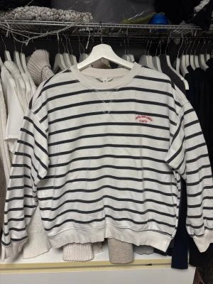 Randig vit och svart sweatshirt - Säljer en vit sweatshirt med svarta horisontella ränder och röd broderad text 'perdu mon coeur à paris' på bröstet.