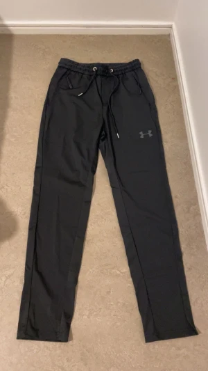 Svarta träningsbyxor från Under Armour - Svarta träningsbyxor från Under Armour med smal passform och elastisk midja med snörning. Byxorna har två sidofickor och en diskret logga på vänster ben. Materialet är lätt och syntetiskt, perfekt för aktiva dagar.