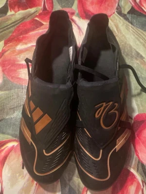 Adidas Predator fotbollsskor svart/guld - Säljer ett par Adidas Predator fotbollsskor i storlek 42. Skorna är svarta med snygga gulddetaljer och har snörning samt mönstrad ovandel. Utrustade med dobbar för gräsplan och tillverkade i syntetmaterial. Perfekt för dig som vill ha grym kontroll på planen.