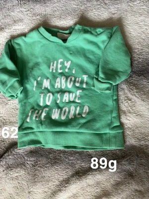 Grön sweatshirt med tryck, stl 62 - Cool grön sweatshirt med vitt tryck 'Hey, I'm about to save the world' framtill. Tröjan har långa ärmar, ribbade muddar och knappar på axeln för enkel påklädning. Perfekt för en liten hjälte!🌸Fint använt skick. Storlek 62