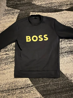 Svart sweatshirt från BOSS med gul text - Svart sweatshirt från BOSS med stor gul BOSS-logga på bröstet. Säljer en Hugo Boss i storlek M. Den är knappt använd, endast två gånger, och är i nyskick utan skador eller fläckar. Passar perfekt för dig som vill ha hög kvalitet till bra pris. Skicka meddelande vid intresse!